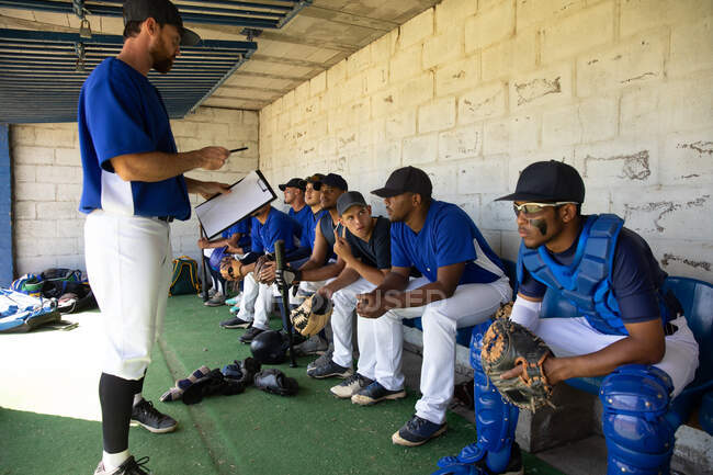 Vue de côté d'un entraîneur de baseball masculin caucasien expliquant un plan de match et donnant des instructions à une équipe multiethnique de joueurs de baseball masculins, se préparant avant un match, assis dans un vestiaire — Photo de stock