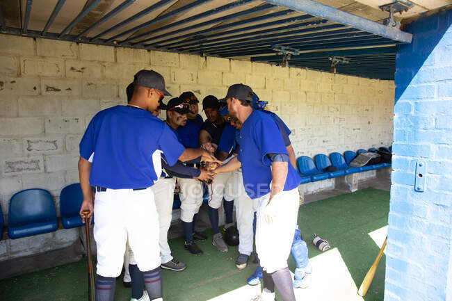 Rückansicht einer multiethnischen Gruppe männlicher Baseballspieler, die sich vor einem Spiel vorbereitet, in einer Umkleidekabine steht, sich kauert, mit der Faust schlägt, miteinander interagiert, sich gegenseitig motiviert — Stockfoto