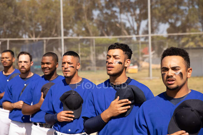 Seitenansicht einer multiethnischen Gruppe männlicher Baseballspieler, die sich vor einem Spiel vorbereiten, in einer Reihe stehen, einer Nationalhymne lauschen und singen, ihre Mützen auf der Brust haltend — Stockfoto