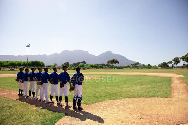 Vista trasera de un grupo multiétnico de jugadores de béisbol masculinos, preparándose antes de un partido, de pie en fila, escuchando un himno nacional, sosteniendo sus gorras en el pecho - foto de stock