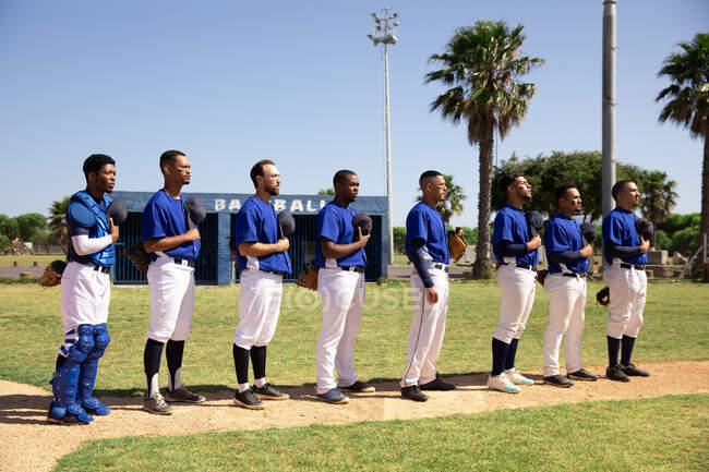 Seitenansicht einer multiethnischen Gruppe männlicher Baseballspieler, die sich vor einem Spiel vorbereiten, in einer Reihe stehen, einer Nationalhymne lauschen und ihre Mützen auf der Brust halten — Stockfoto