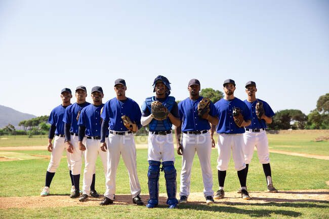 Vista frontale di un gruppo multietnico di giocatori di baseball maschi, che si preparano prima di una partita, in piedi uno accanto all'altro su un campo da baseball, tenendo i guanti e un casco da baseball — Foto stock