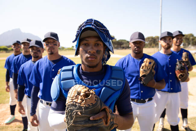 Vista frontale di un gruppo multietnico di giocatori di baseball maschi, che si preparano prima di una partita, in piedi uno accanto all'altro su un campo da baseball, tenendo i guanti e un casco da baseball — Foto stock