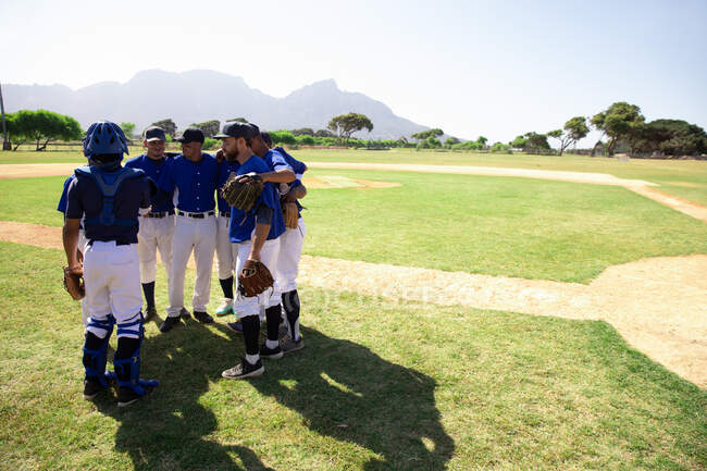 Seitenansicht einer multiethnischen Mannschaft männlicher Baseballspieler, die sich vor einem Spiel in einem Gedränge auf einem Baseballfeld vorbereitet und ihrem Kapitän zuhört, der ihnen Anweisungen gibt, an einem sonnigen Tag — Stockfoto