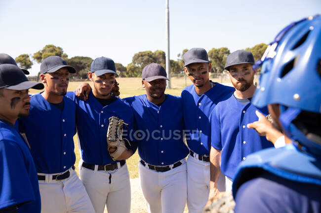 Вид спереди на многонациональную команду бейсболистов-мужчин, готовящихся перед игрой, в кучке на бейсбольном поле, слушающих, как их капитан дает им указания, в солнечный день — стоковое фото