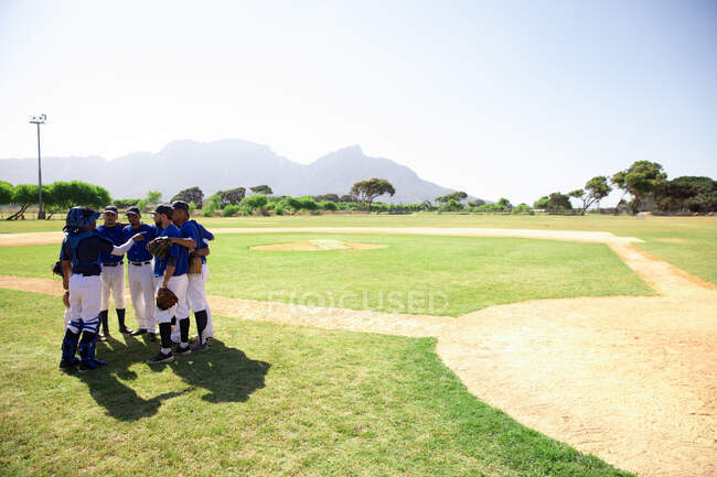Vista lateral de un equipo multiétnico de jugadores de béisbol que se preparan antes de un partido, en un grupo en un campo de béisbol, escuchando a su capitán darles instrucciones, en un día soleado - foto de stock