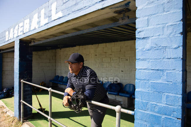 Vue latérale d'un arbitre de baseball mixte senior, se préparant avant un match, debout dans un vestiaire, appuyé sur une barrière, tenant un casque — Photo de stock
