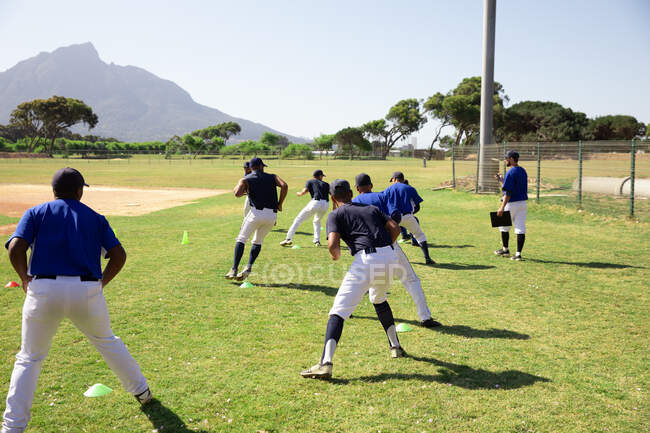 Vista posteriore di un gruppo multietnico di giocatori di baseball maschili, allenamento con il loro allenatore in un campo da gioco, allenamento, corsa, in una giornata di sole — Foto stock