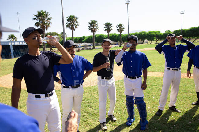 Side view of a team of multi-ethnic male baseball players, preparing before a game, resting, drinking water and talking on a sunny day — Stock Photo