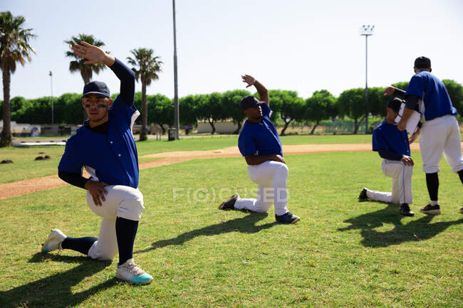 Vue latérale d'un groupe multiethnique de joueurs de baseball masculins, se préparant avant un match, s'entraînant, s'agenouillant et s'étirant d'affilée, par une journée ensoleillée — Photo de stock