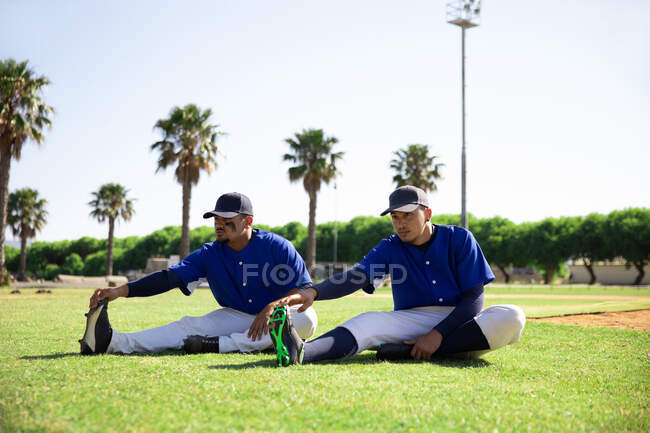 Vue latérale de deux joueurs de baseball de race mixte assis, étirant leurs jambes, lors d'une séance d'entraînement sur un terrain de jeu par une journée ensoleillée d'affilée, par une journée ensoleillée — Photo de stock