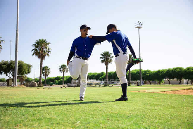 Вид сбоку кавказца и смешанной расы бейсболистов, опирающихся друг на друга на плечи, стоящих на одной ноге, держащих и растягивающих другую ногу, во время тренировки на игровом поле в солнечный день — стоковое фото