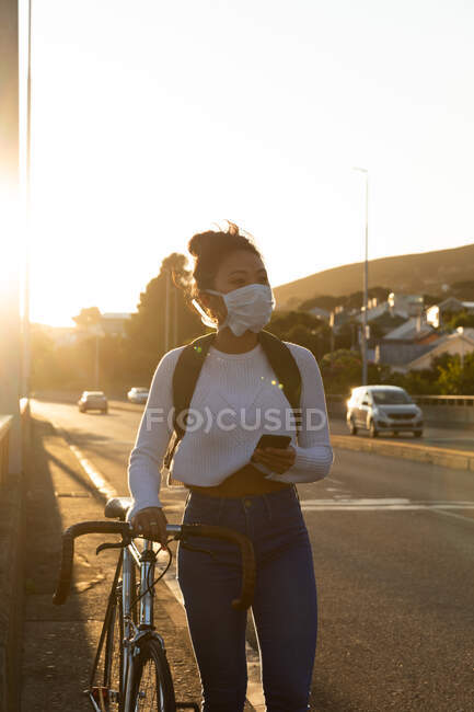 Vista frontal de una mujer de raza mixta con el pelo largo y oscuro por las calles de la ciudad durante el día, con una máscara facial contra la contaminación del aire y el coronavirus, caminando con su bicicleta y usando un teléfono inteligente - foto de stock