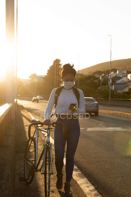 Вид спереду змішаної раси жінка з довгим темним волоссям на вулицях міста протягом дня, вдягнена маска для обличчя проти забруднення повітря та коронавірусу, що йде з велосипедом та використовує смартфон — стокове фото