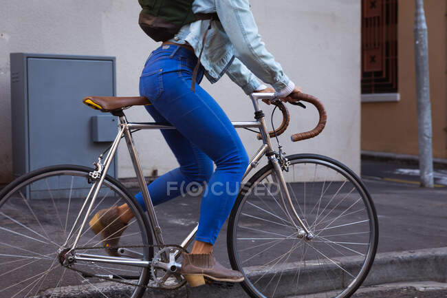 Seitenansicht Unterteil einer Frau, die tagsüber in den Straßen der Stadt unterwegs ist, Skinny Jeans und braune Stiefel mit Rucksack trägt und auf ihrem Fahrrad in einer Stadtstraße mit Gebäude im Hintergrund unterwegs ist. — Stockfoto