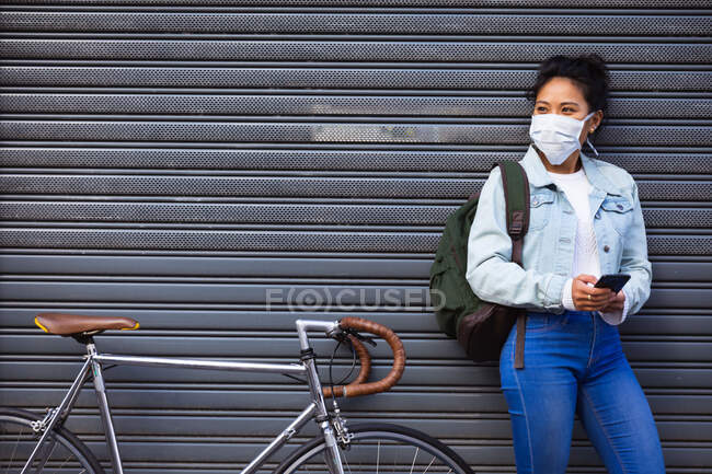 Вид спереди на женщину смешанной расы с длинными темными волосами на улицах города в течение дня, одетую в маску против загрязнения воздуха и коронавируса, стоящую рядом с велосипедом и использующую смартфон — стоковое фото