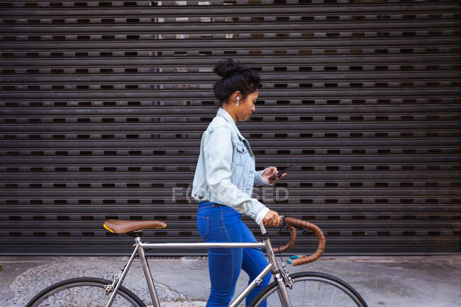 Vue latérale d'une femme métisse aux longs cheveux foncés dans les rues de la ville pendant la journée, marchant à vélo et utilisant un smartphone, écouteurs allumés avec volets gris en arrière-plan. — Photo de stock