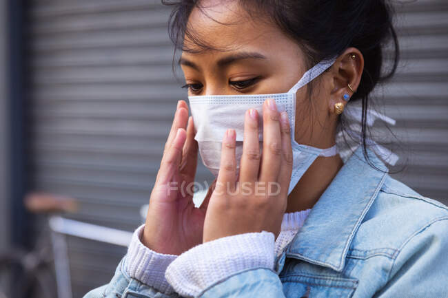 Side view close up of a mixed race woman with dark hair out and about in the city streets during the day, wearing a face mask against air pollution and coronavirus, standing with hands to her mouth with her bicycle in the background. — Stock Photo