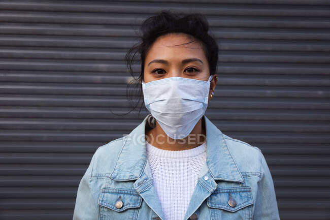 Portrait d'une femme métisse aux cheveux longs et foncés dans les rues de la ville pendant la journée, portant un masque facial contre la pollution atmosphérique et le coronavirus, debout et regardant la caméra avec un mur gris en arrière-plan. — Photo de stock