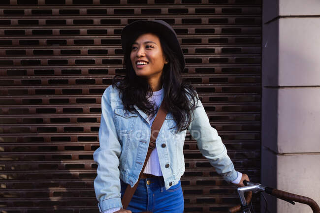 Vue de face d'une heureuse femme métisse aux longs cheveux foncés dans les rues de la ville pendant la journée, vêtue d'un chapeau et d'une veste en denim, appuyée sur son vélo souriant avec un mur en arrière-plan. — Photo de stock