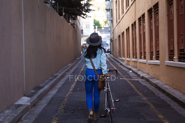 Vue arrière d'une heureuse femme métissée aux longs cheveux foncés dans les rues de la ville pendant la journée, portant un chapeau, un jean et une veste en denim, marchant avec son vélo avec des bâtiments en arrière-plan. — Photo de stock