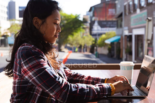 Vue latérale d'une femme métisse aux longs cheveux foncés dans les rues de la ville pendant la journée, assise dans un café travaillant sur un ordinateur portable avec des bâtiments en arrière-plan. — Photo de stock