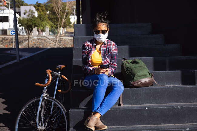 Vue de face d'une femme métisse aux cheveux foncés dans les rues de la ville pendant la journée, portant des lunettes de soleil et un masque facial contre la pollution atmosphérique et le coronavirus, assise sur des marches à l'aide d'un smartphone — Photo de stock