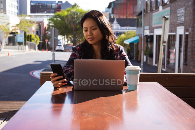 Vue de face d'une femme métisse aux longs cheveux foncés assise à une table dans un café pendant la journée, travaillant sur un ordinateur portable à l'aide d'un smartphone avec des bâtiments en arrière-plan. — Photo de stock