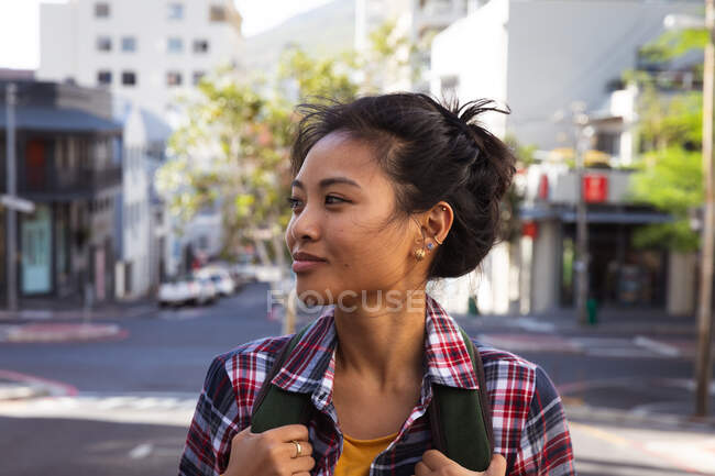 Вид збоку крупним планом щаслива змішана жінка з довгим темним волоссям на вулицях міста протягом дня, несучи рюкзак, одягнений в перевірену сорочку, посміхаючись з будівлями на задньому плані . — стокове фото