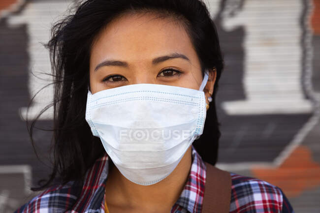 Portrait d'une femme métisse aux cheveux longs et foncés dans les rues de la ville pendant la journée, portant un masque facial contre la pollution de l'air et le coronavirus, debout regardant la caméra avec un mur en arrière-plan. — Photo de stock