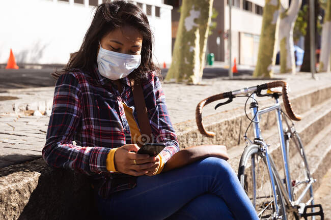 Vue latérale d'une femme métisse aux cheveux foncés dans les rues de la ville pendant la journée, portant des lunettes de soleil et un masque facial contre la pollution atmosphérique et le coronavirus, assise sur des marches à l'aide d'un smartphone — Photo de stock