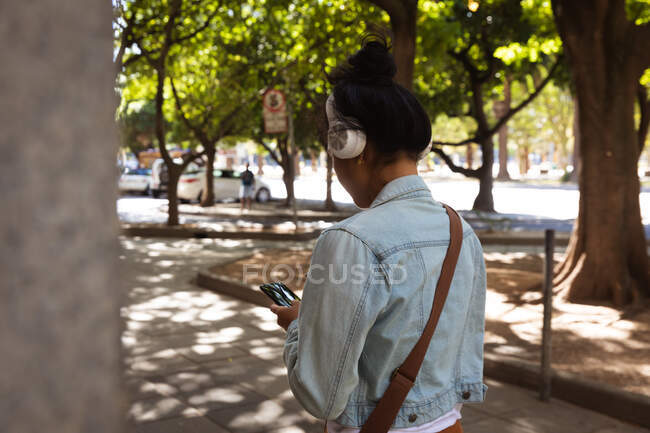 Vue arrière d'une femme métisse aux longs cheveux foncés dans les rues de la ville pendant la journée, utilisant un smartphone, portant un casque, une veste en denim et se promenant dans une rue de la ville avec des arbres et des voitures en arrière-plan. — Photo de stock