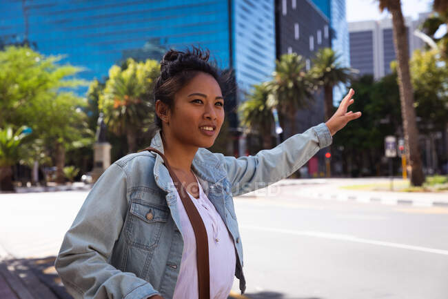 Вид сбоку счастливой смешанной расовой женщины с длинными темными волосами на улицах города в течение дня, стоящей на городской улице, поднимающей руку, чтобы остановить такси, в джинсовой куртке, улыбающейся со зданиями на заднем плане. — стоковое фото