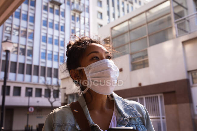 Niedrige Seitenansicht einer Mischlingsfrau mit langen dunklen Haaren, die tagsüber auf den Straßen der Stadt unterwegs ist und eine Gesichtsmaske gegen Luftverschmutzung und Coronavirus trägt, mit Gebäuden im Hintergrund. — Stockfoto