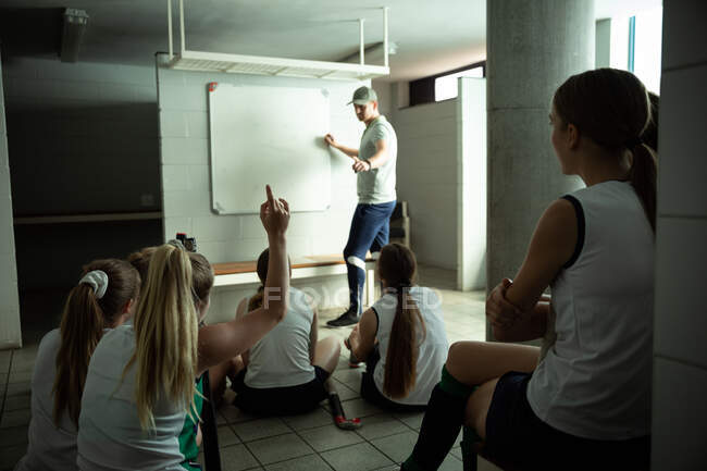 Side view of a Caucasian male field hockey coach interacting with a group of female Caucasian field hockey players, sitting in a changing room, showing them a game plan, writing on a whiteboard, with one of the players raising her hand — Stock Photo