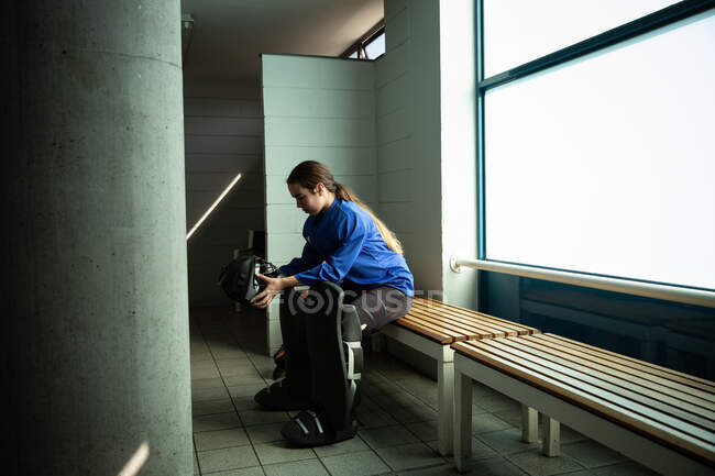Vue latérale d'une joueuse de hockey sur gazon caucasienne se préparant avant un match, assise dans un vestiaire, portant un casque de hockey — Photo de stock