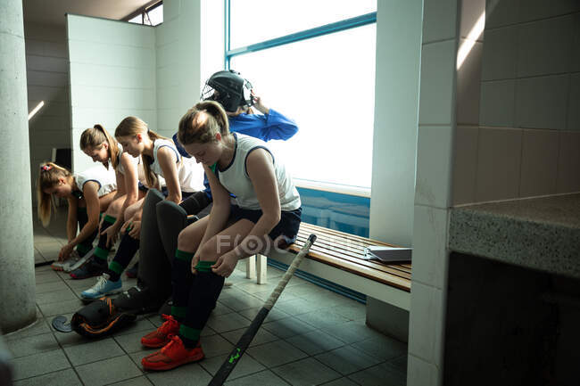 Vue latérale d'un groupe de joueuses de hockey sur gazon caucasiennes se préparant avant un match, assises dans un vestiaire, portant leurs chaussures et guêtres — Photo de stock