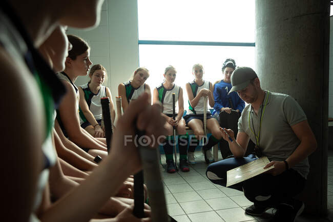 Vista lateral de um treinador de hóquei em campo caucasiano interagindo com um grupo de jogadoras de hóquei em campo caucasianas, sentadas em um vestiário, mostrando-lhes um plano de jogo — Fotografia de Stock