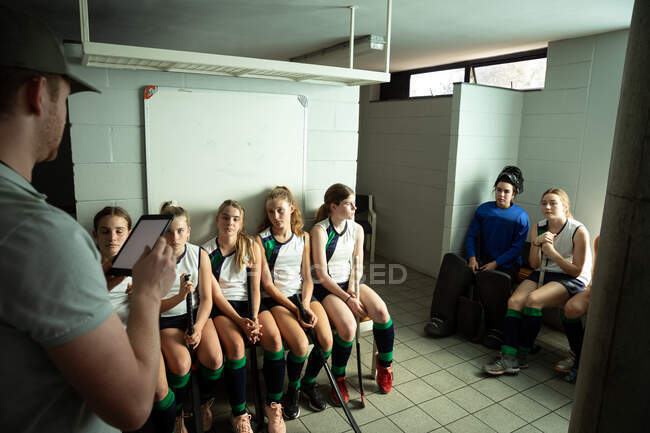 Seitenansicht eines kaukasischen Hockeytrainers, der mit einer Gruppe kaukasischer Hockeyspielerinnen interagiert, in einer Umkleidekabine sitzt und ein digitales Tablet in der Hand hält — Stockfoto