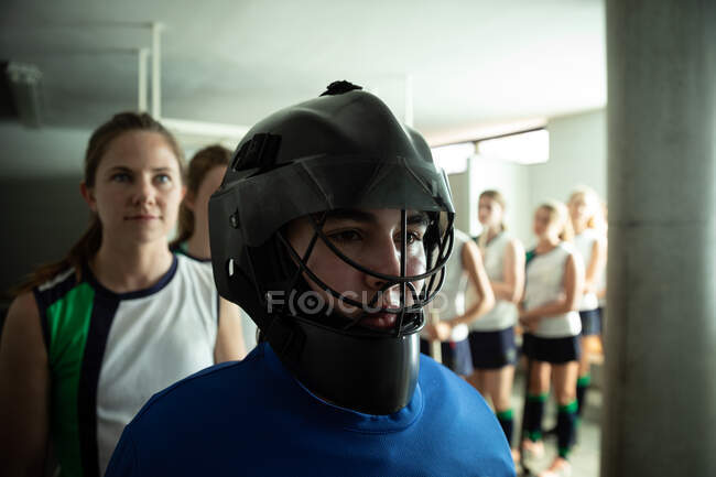 Side view close up of a Caucasian female field hockey players, preparing before a game, standing in a changing room, wearing a hockey helmet, with her teammates standing in a row behind her — Stock Photo