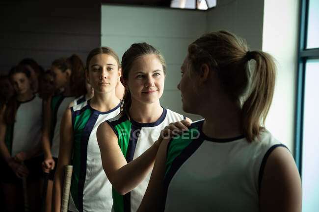 Vorderansicht einer Gruppe kaukasischer Hockeyspielerinnen, die sich vor einem Spiel vorbereiten, in einer Umkleidekabine stehend, legt eine ihre Hand auf die Schulter ihrer Teamkollegin — Stockfoto
