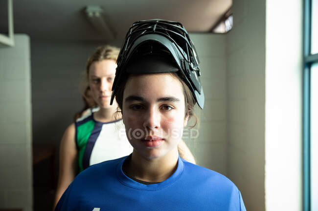 Retrato de una jugadora de hockey caucásica, preparándose antes de un partido, de pie en un vestuario, usando un casco de hockey, con sus compañeras de equipo de pie en una fila detrás de ella - foto de stock