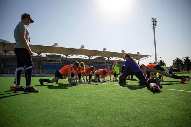 Side view of a group of female Caucasian field hockey players and their Caucasian male field hockey coach, training before a game, working out on a field hockey pitch, doing exercises, stretching their legs, on a sunny day — Stock Photo