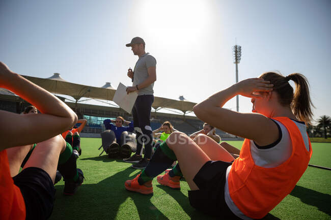 Side view of a group of female Caucasian field hockey players and their Caucasian field hockey coach, training before a game, working out on a field hockey pitch, exercising, doing sit ups, on a sunny day — Stock Photo