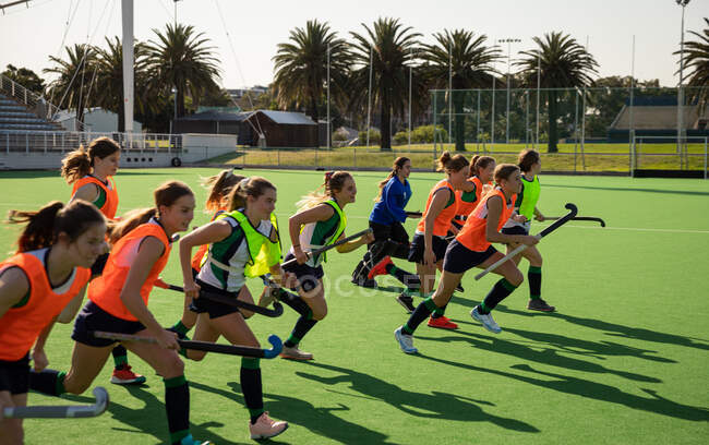 Side view of a group of female Caucasian field hockey players training before a game, working out on a field hockey pitch, doing exercises, running, holding hockey sticks on a sunny day — Stock Photo