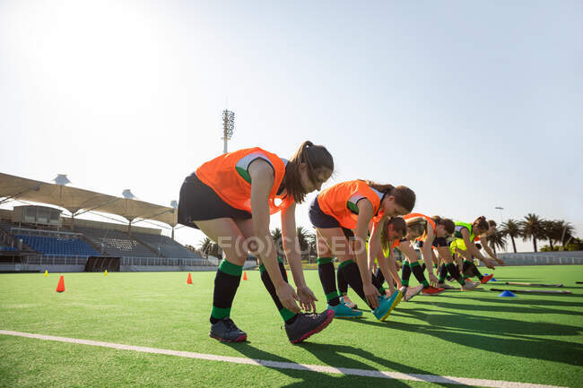 Seitenansicht einer Gruppe kaukasischer Hockeyspielerinnen, die vor einem Spiel trainieren, auf einem Feldhockeyfeld trainieren, Übungen machen, die Beine strecken, sich an einem sonnigen Tag zu den Füßen bücken — Stockfoto