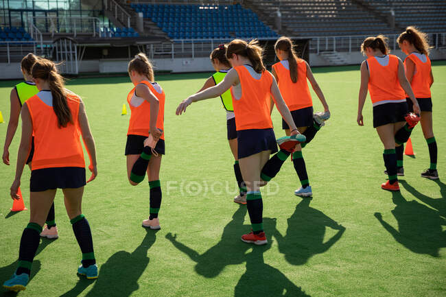Side view of a group of female Caucasian field hockey players, training before a game, working out on a field hockey pitch, doing exercises, stretching their legs, holding their feet behind their back on a sunny day — Stock Photo