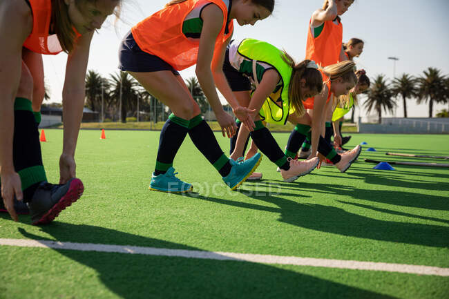 Side view of a group of female Caucasian field hockey players, training before a game, working out on a field hockey pitch, doing exercises, stretching their legs, bending down to their feet on a sunny day — Stock Photo