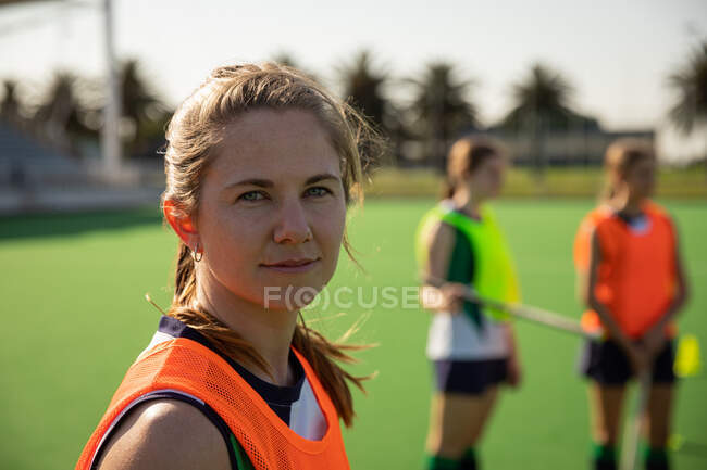 Portrait of a Caucasian female field hockey player, training before a game, working out on a hockey pitch, looking at camera, with her teammates in the background on a sunny day — Stock Photo