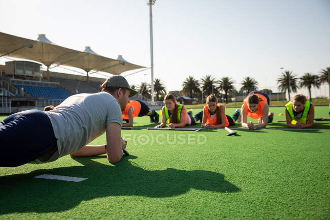 Front view of a group of female Caucasian field hockey players and their Caucasian male field hockey coach, training before a game, working out on a field hockey pitch, doing planks, on a sunny day — Stock Photo